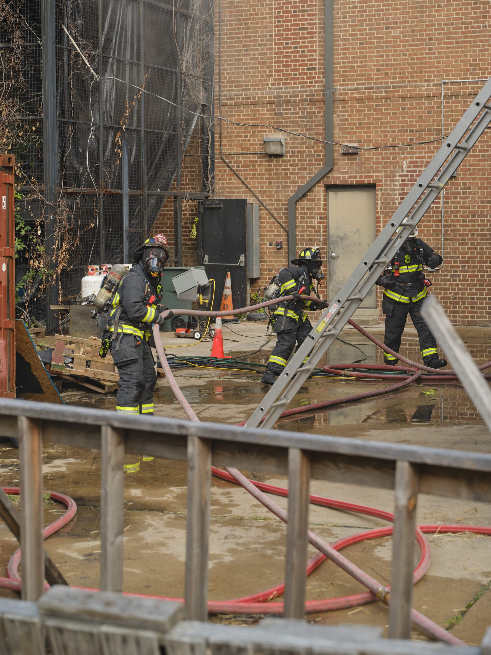 Feuerwehrleute in Helmen arbeiten daran, ein Gebäudefeuer zu löschen, umgeben von Ausrüstung und einem Metallzaun.