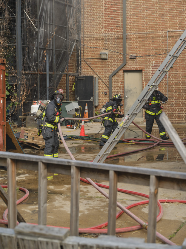 Feuerwehrleute in Helmen arbeiten daran, ein Gebäudefeuer zu löschen, umgeben von Ausrüstung und einem Metallzaun.