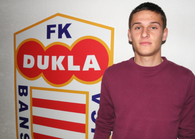 Ein Mann in einem rotbraunen T-Shirt steht vor einer weißen Wand mit einem Schild, auf dem "FK Dukla" steht und zeigt einen entschlossenen Gesichtsausdruck.