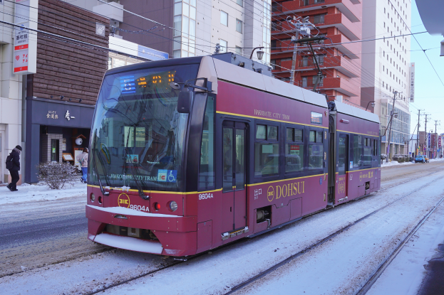 Ein roter und gelber Zug fährt auf einer verschneiten Straße mit hohen Gebäuden, Strommästen, Schildern, Pflanzen und einer Gruppe von Menschen unter einem bewölktem Himmel.