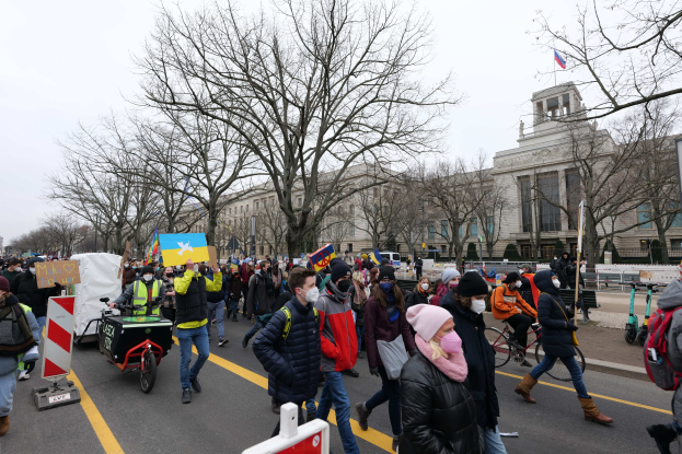 A large protest march with people walking down a street in Washington, D.C., some holding signs and others riding bicycles, with trees, signboards, and a clear blue sky in the background.