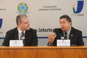 Zwei Männer in Anzügen sitzen an einem Tisch mit zwei Namensschildern und zwei Mikrofonen, mit Logos und Text im Hintergrund.