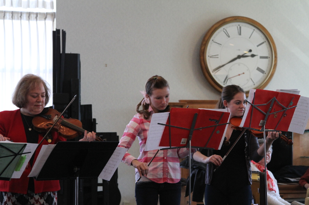 Drei Frauen, die auf der Bühne Geigen spielen, mit Notenpulten davor, einem Publikum im Vordergrund, einer Uhr an einer weißen Wand im Hintergrund und einem weißen Vorhang in der linken Ecke.