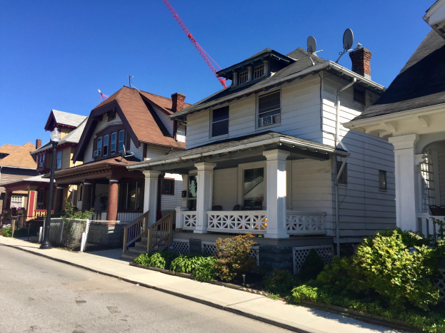 Eine Reihe von Häusern mit architektonischen Details wie Fenstern, Säulen, Geländern, Treppen, Dächern, featuring einen Baukran auf einem Dach, einen Straßen, Pflanzen, einen Zaun, einen Weg, Bäume und einen bewölkten Himmel.