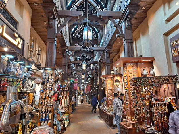 Ein belebter Dubai-Basar mit Menschen, die zwischen Regalen mit Waren hindurchgehen, beleuchtet von Deckenlampen und mit Texttafeln an den Wänden.