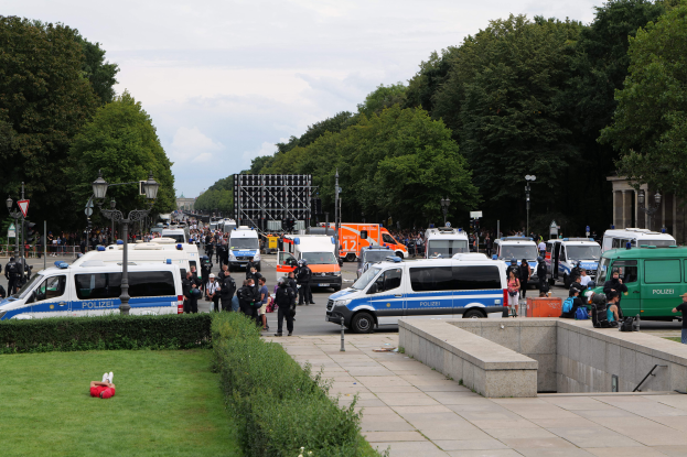 Eine große Gruppe von Polizeiwagen am Straßenrand in Berlin geparkt, mit vielen Menschen um die Fahrzeuge herum, einige mit Taschen, und Laternenpfähle, Bäume und ein Gebäude im Hintergrund unter einem bewölkten Himmel.