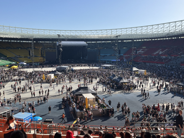 A large crowd stands in front of a stadium with railings, tents, and stage equipment in the foreground, while a lit stage with trusses is visible in the background under a clear sky.