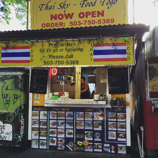 Ein Schaufenster mit verschiedenen Gegenständen wie Getränkedosen, Speisekarten, einer Uhr, Gewürzen in Plastikbehältern, Kondimente, Werbetafeln und Namensschildern unter einem Himmel-Hintergrund.