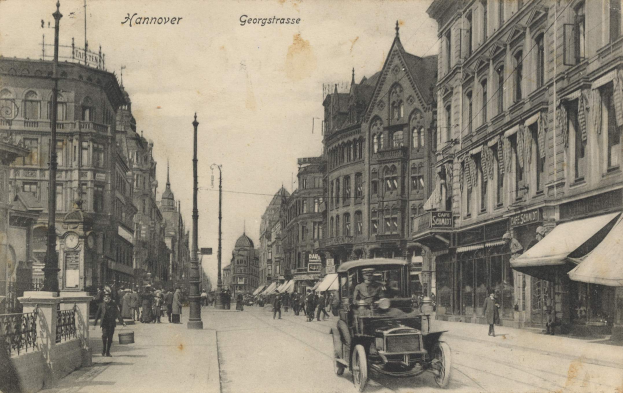 Ein altes Schwarz-Weiß-Foto einer Straße in Hannover, Deutschland, das Fahrzeuge auf der Straße, Menschen auf dem Gehweg, Gebäude mit Fenstern auf beiden Seiten, Masten, eine Uhr und den Himmel im Hintergrund zeigt.