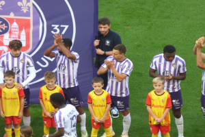 Eine Gruppe junger Jungs steht auf einem Fußballfeld, klatscht in die Hände, mit einem Banner und Logos im Hintergrund und saftigem grünem Gras darunter.