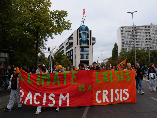 Group of people walking down a street holding a "Climate Crisis is a Crisis" banner, with trees, light poles, and buildings lining the street.