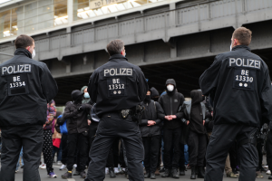 Eine Gruppe uniformierter Polizisten steht vor einer Menge von Menschen in schwarzen Uniformen und Masken mit einer Brücke und einem Gebäude im Hintergrund während einer Demonstration in einer Stadt.