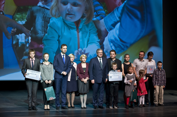 Eine Gruppe von Menschen auf einer Bühne mit Taschen und Urkunden, mit einem Bildschirm im Hintergrund, der ein Kind und andere zeigt.