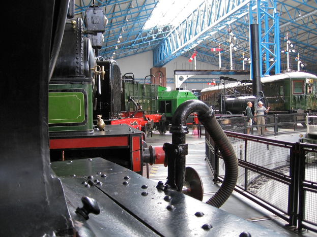 Mehrere Zugmaschinen stehen in einem Lagerhaus mit zahlreichen Eisenstangen darüber, einem Zaun auf der rechten Seite, einem Zug auf dem Bahngleis neben dem Zaun und zwei Personen in der Mitte nahe dem Zaun.