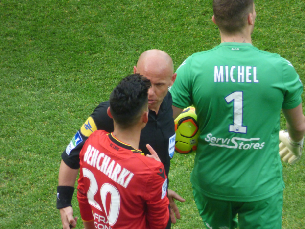 Referee speaking with two soccer players on a grass-covered field, one player holding a ball.