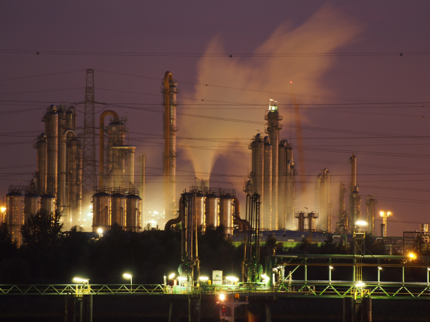 Ölraffinerie bei Nacht mit aufsteigendem Rauch, beleuchtet von umliegenden Lichtern, vor einem sichtbaren Himmel, mit einem Wasserzeichen.