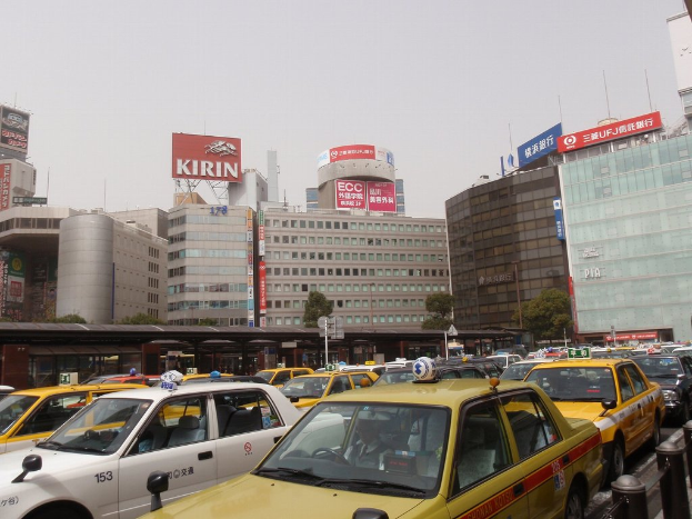 Stadtansicht mit Fahrzeugen auf der Straße, Gebäuden, Bäumen, Pfählen mit Schildern und Text auf den Schildern, einem Zug im Hintergrund und einem Geländer unten rechts.