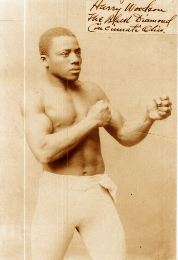 Ein Schwarz-Weiß-Foto eines Mannes im Boxring mit Handschuhen und Shorts, der mit konzentrierter Miene und entschlossener Haltung steht.