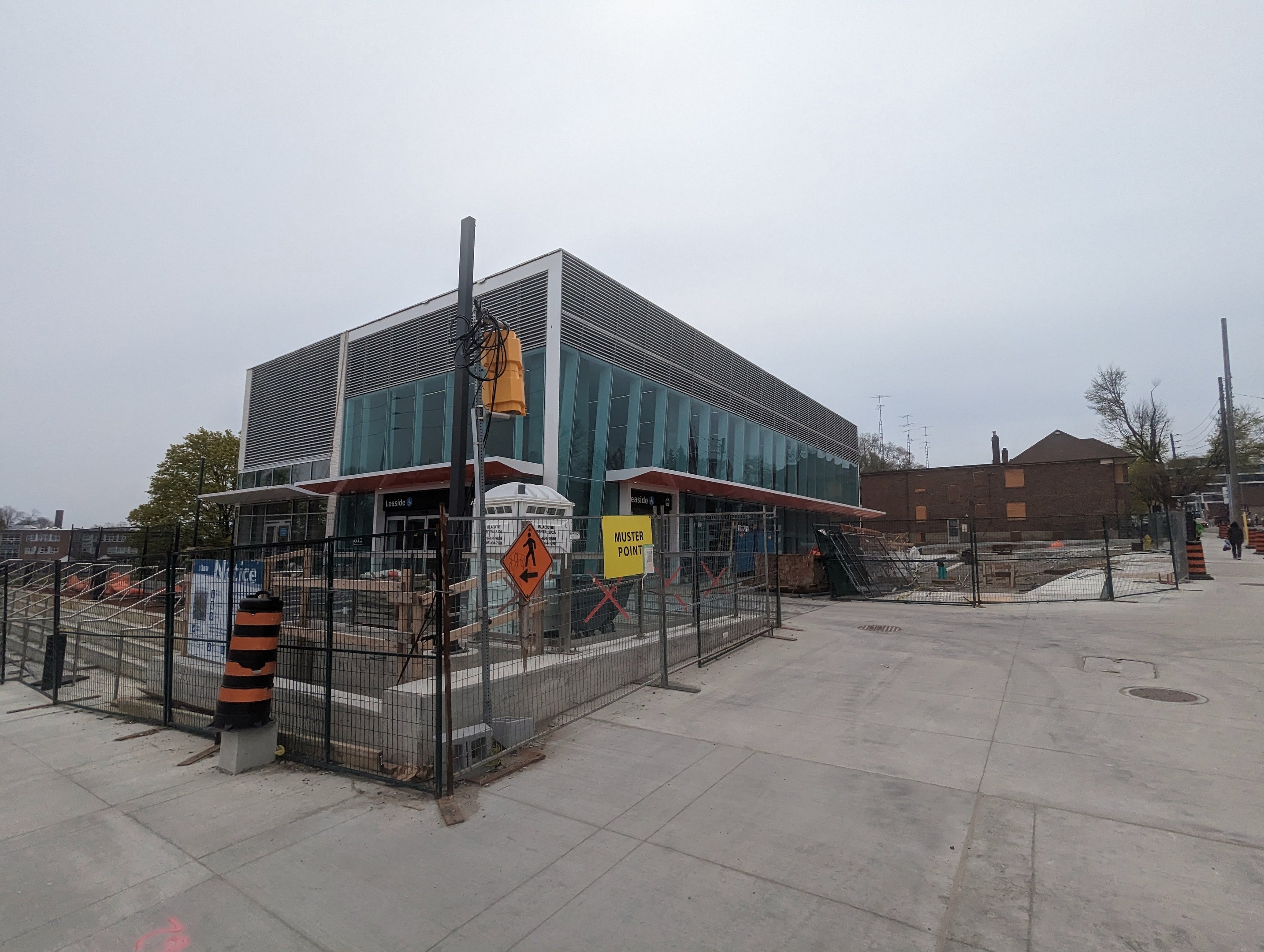 Large multi-story school building with numerous windows, surrounded by a metal fence, trees, signboards, and a few people and vehicles; cloudy sky overhead.