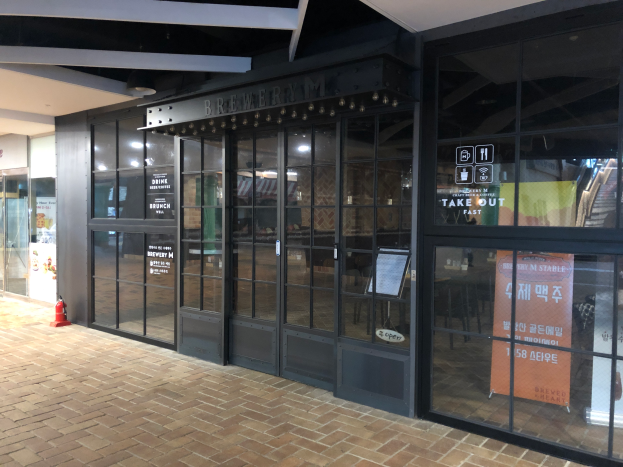 Der Eingang eines Gebäudes mit Glaswänden und -türen, ein Schild mit der Aufschrift "Brewery M Stable", ein Feuerhydrant auf dem Boden und Deckenleuchten, die den Bereich beleuchten.