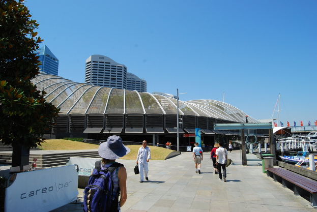 Menschen, die auf einem Gehweg mit Mülltonnen, Bänken, Holzlatten, Metallstangen auf der rechten Seite, Pfählen, Bäumen und einem Metallstangenzaun mit Bannern auf der linken Seite sowie Gebäuden im Hintergrund gehen.