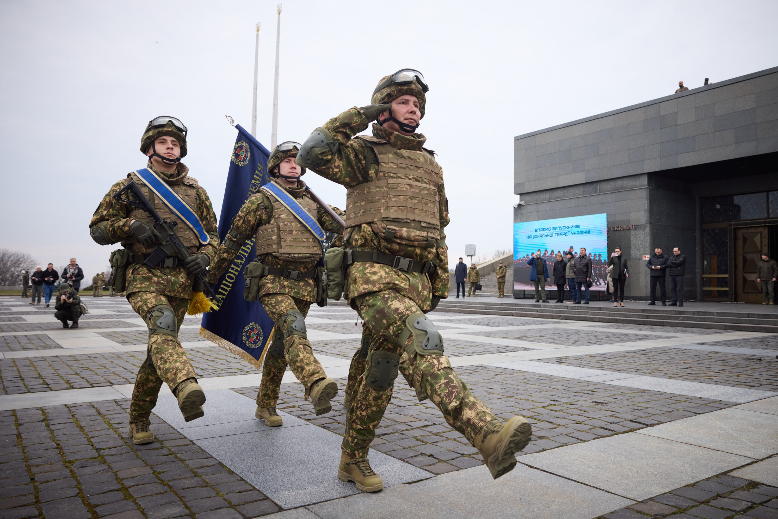 Group of Ukrainian soldiers in uniform marching down a street, carrying guns and flags, with onlookers and a building in the background.