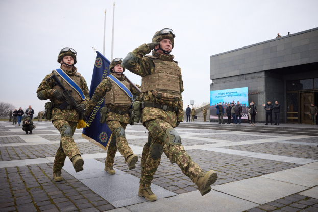 Group of Ukrainian soldiers in uniform marching down a street, carrying guns and flags, with onlookers and a building in the background.