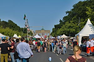 Eine Menschenmenge, die eine Straße entlanggeht, gesäumt von Zelten, Fahrzeugen und Bäumen, mit einem Bogen und einem klaren blauen Himmel im Hintergrund und Polen mit Fahnen auf der linken Seite, die wahrscheinlich das Oktoberfest in München, Deutschland, darstellen.