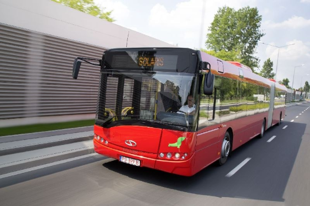 Ein roter Bus fährt auf einer Straße neben einem Gebäude entlang, mit einer Person im Inneren, neben einer Wand, Bäumen, Laternen und einem bewölkten Himmel im Hintergrund.