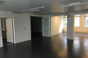 Ein leeres Büro mit schwarzen Böden, weißen Wänden, elektrischen Lampen, Kabeln, Türen, Schränken und Fenstern mit Blick auf Gebäude und Bäume in Amsterdam.