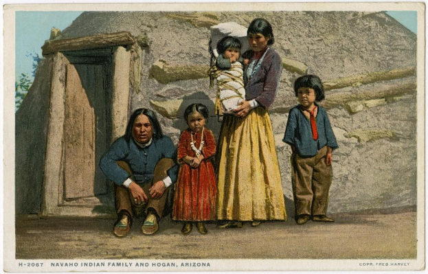 Old postcard of a Navajo family posing in front of a house with a door, surrounded by trees under a clear blue sky, with text reading "Navajo Indian Family and Hogan, Arizona" at the bottom.