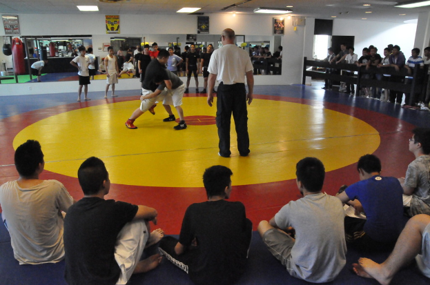 Zwei Menschen kämpfen auf einer Matte, während ein Mann in einem weißen T-Shirt daneben steht; Zuschauer schauen von hinter einer Absperrung zu, einige sitzen auf dem Boden, mit Spiegeln, Boxsäcken und Postern im Hintergrund.