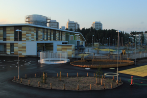 Leerer Parkplatz mit einem Spielplatz in der Mitte, umgeben von Gebäuden, Straßenlaternen, Straßenlaternen, Bäumen und einem klaren blauen Himmel.