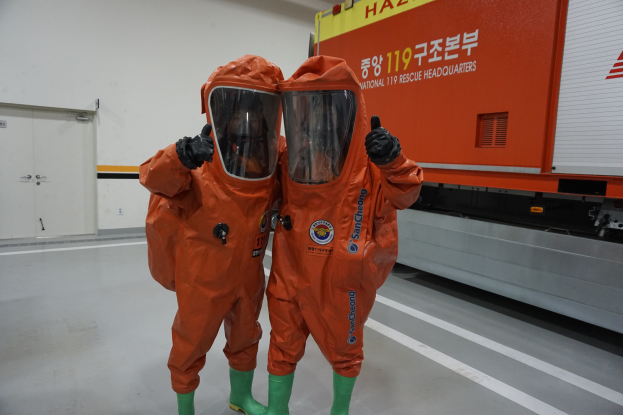Zwei Personen in orangefarbenen Schutzanzügen stehen nebeneinander vor einem Lastwagen, mit einer Wand und Türen im Hintergrund, was darauf hinweist, dass sie Teil des Nationalen Einsatzteams sind.