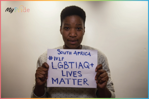 A woman in a white shirt stands determinedly in front of a wall, holding a sign that reads "LGBTQ+ Lives Matter" in bold black lettering.
