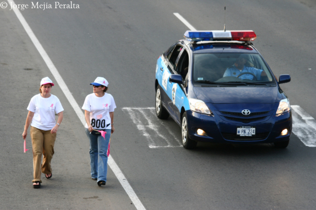 Zwei Frauen gehen auf einer Straße, eine hölt ein rosa Papier und trägt eine rosa und weiße Kappe, die andere hölt ein rosa Papier und trägt eine blaue und weiße Kappe, mit einem patrouillierenden Polizeiwagen und seinem Fahrer auf der rechten Seite.