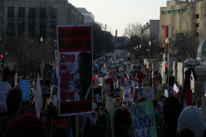 Menschen mit Plakaten auf einer Straße, mit Gebäuden, Bäumen, Laternen und Himmel im Hintergrund.