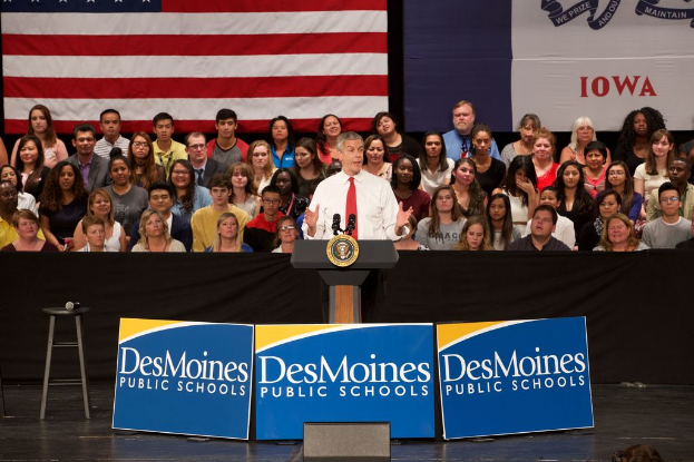 Ein Mann in einem weißen Hemd und roter Krawatte steht an einem Podium mit drei Tafeln mit Text, einem Hocker mit Mikrofon und einer sitzenden Menge im Hintergrund, mit einer Flagge und zusätzlichem Text.