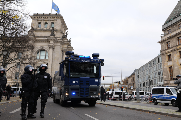 Eine Gruppe von Polizisten in Helmen und mit Waffen steht vor einem Gebäude mit Fenstern, Säulen, Bögen und einer Flagge, mit Fahrzeugen auf der Straße, Bäumen, Laternenmasten und einem bewölkten Himmel im Hintergrund.
