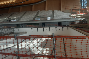 Eine große Aula im Bau mit Gerüsten, einem Sicherheitsnetz unten und verschiedenen Gegenständen wie Stühlen, Treppen, Geländern und Lampen im Hintergrund, mit Fenstern auf der rechten Seite.