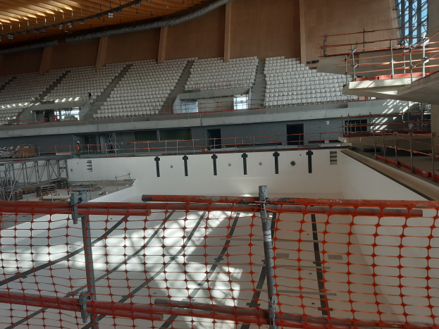 Eine große Aula im Bau mit Gerüsten, einem Sicherheitsnetz unten und verschiedenen Gegenständen wie Stühlen, Treppen, Geländern und Lampen im Hintergrund, mit Fenstern auf der rechten Seite.