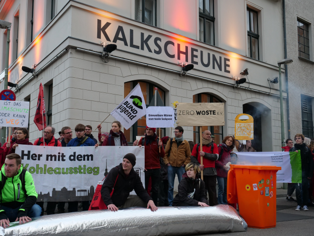 Eine Gruppe von Menschen mit Protestschildern vor einem Gebäude stehend, während andere auf einem Objekt im Vordergrund sitzen und ein Müllcontainer rechts zu sehen ist, mit Gebäuden und Schildern im Hintergrund.