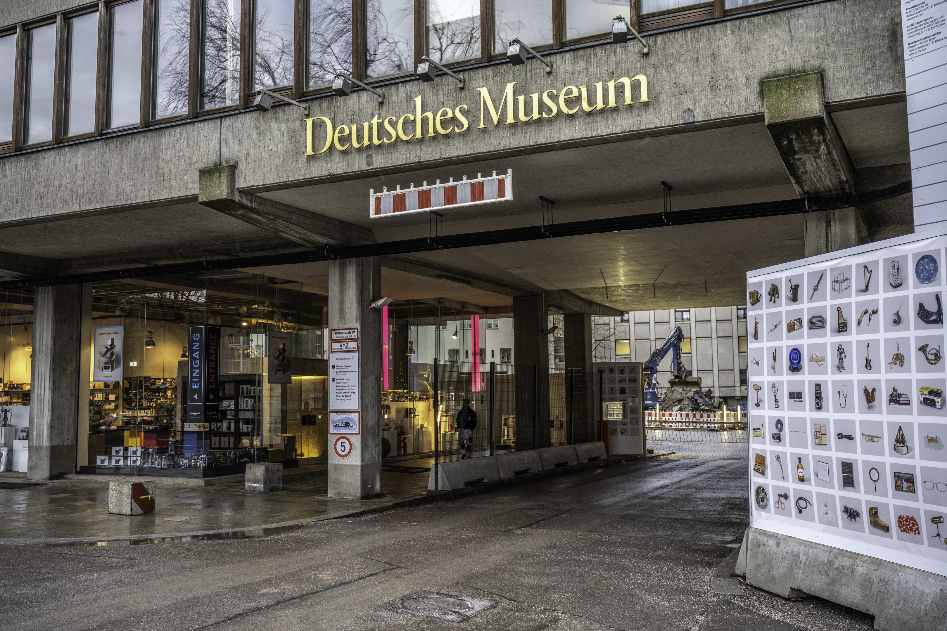 Außensicht des Deutschen Museums in Berlin, das eine große Glasgebäude mit Säulen, ein Namensschild, eine Informationsanzeigetafel und umgebende Gebäude, Bäume und Himmel zeigt.