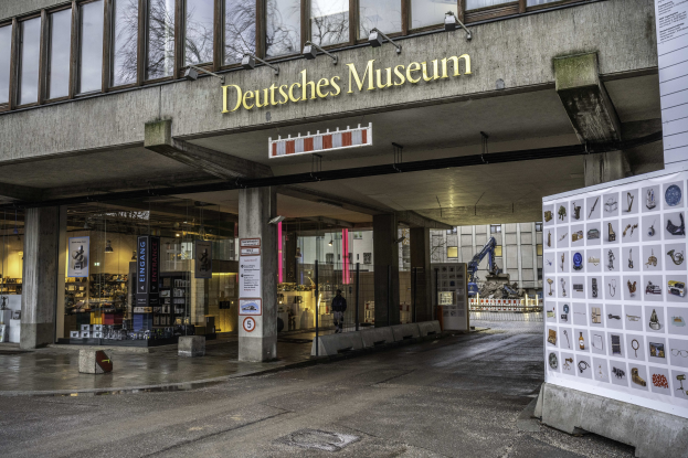 Außensicht des Deutschen Museums in Berlin, das eine große Glasgebäude mit Säulen, ein Namensschild, eine Informationsanzeigetafel und umgebende Gebäude, Bäume und Himmel zeigt.