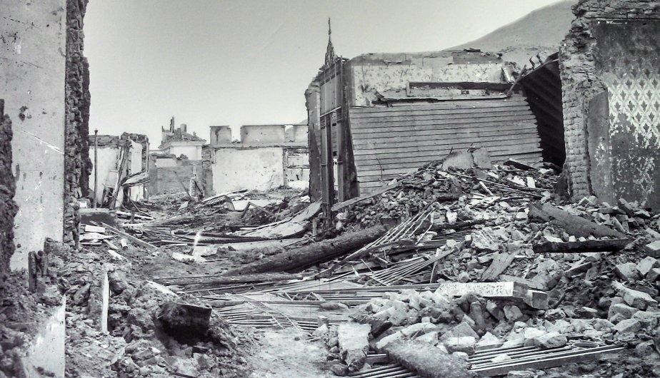 Ein Schwarz-Weiß-Foto eines schwer beschädigten Gebäudes auf einer Straße, umgeben von Trümmern wie eingestürzten Wänden, Holzstöcken und Steinen, mit einem Hügel und Himmel im Hintergrund.