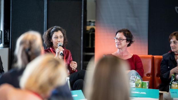 Eine Gruppe von Frauen, die vorne sitzt, wobei eine links eine rote Kleidung trägt und ein Mikrofon hält, und im Hintergrund eine rollende Banner und eine schwarze Decke zu sehen ist.