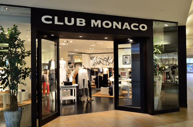 Außenansicht eines Club Monaco Ladens in einem Einkaufszentrum mit Glaswänden, Schaufensterpuppen in Kleidern, Auslagetischen mit Artikeln, einem Topf mit einer Pflanze und Deckenbeleuchtung.