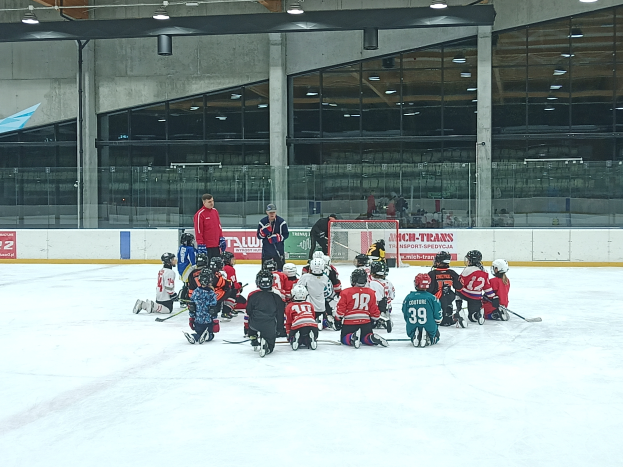 Eine Gruppe von Kindern in Helmen und mit Hockey-Schlägern sitzt auf einer Eisbahn, mit einer Wand aus Glas und Säulen, Deckenlampen und Texttafeln im Hintergrund.