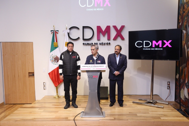 Drei Männer stehen an einem Podium mit einem Mikrofon während einer Pressekonferenz in Mexiko, mit Fahnen, einer Tür und einer Wand mit Text im Hintergrund.