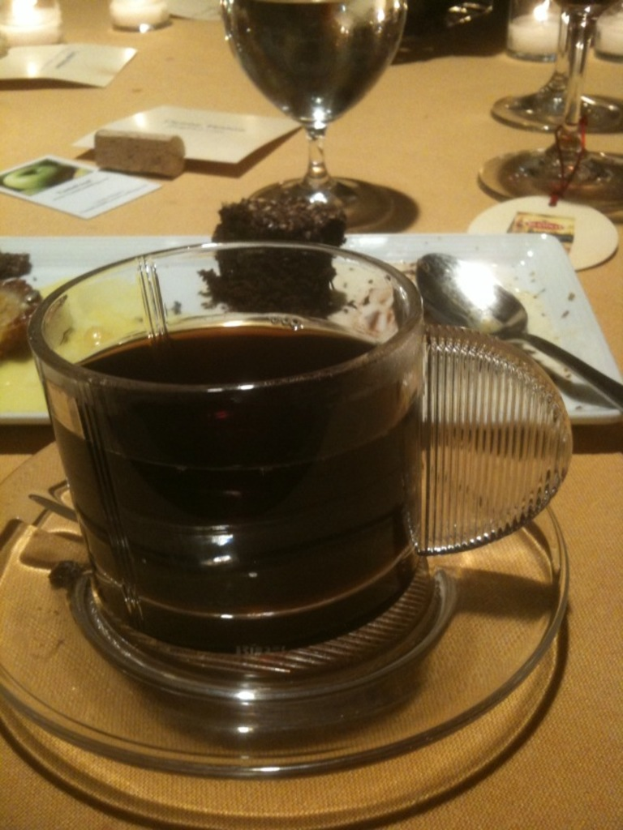 Ein Tisch mit Tasse, Untertasse, Tee, Teller, Löffel, Brownie, Wein, Kerze und zusätzlichem Essen.